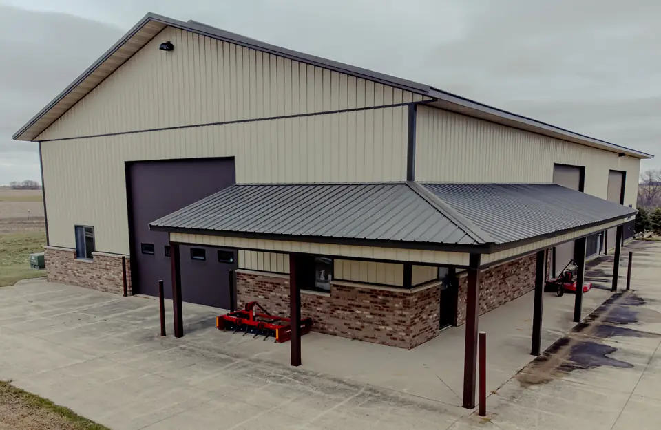 A finished steel-sided post-frame shop with a large overhead door and covered porch beside a farm field